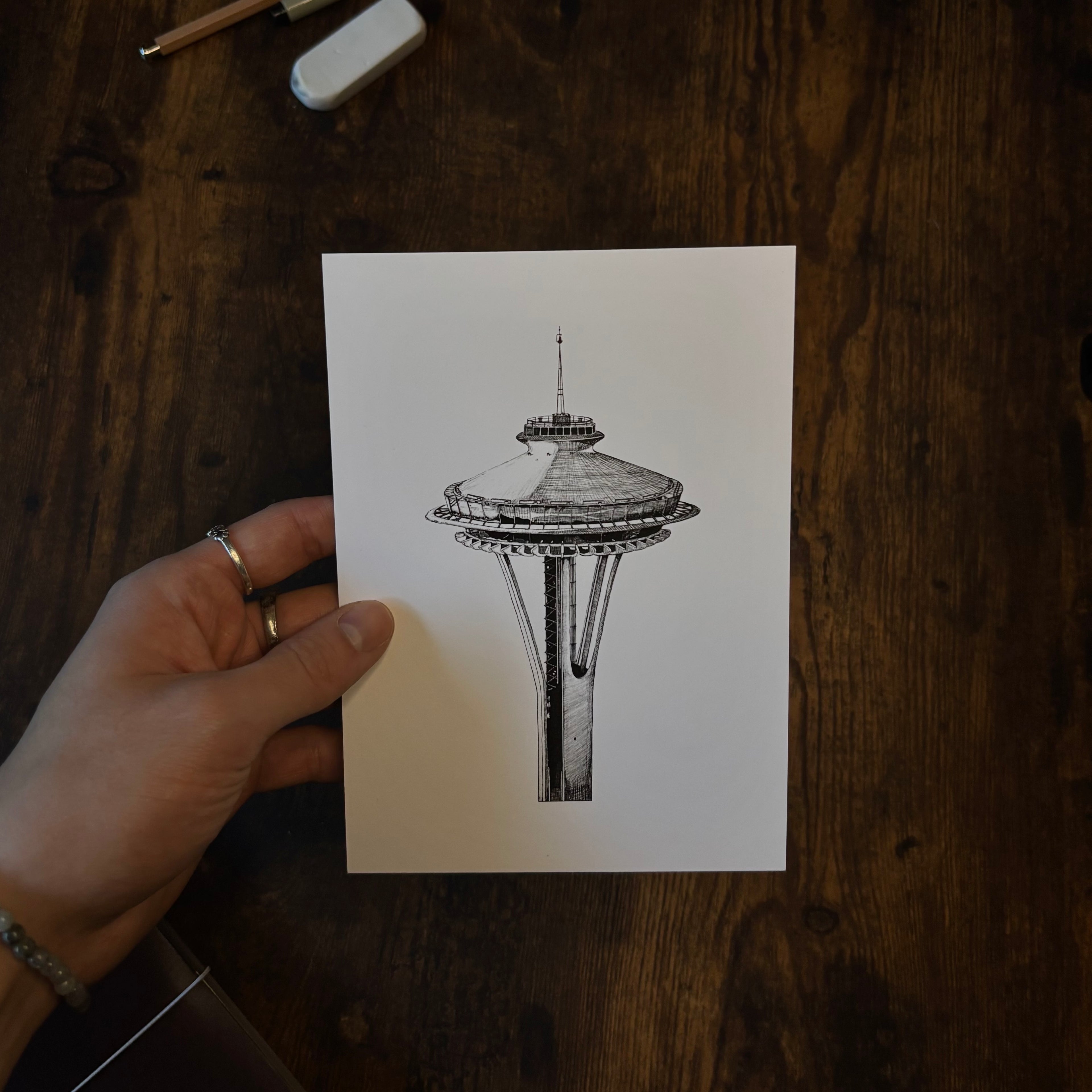 Hand holding a black and white drawing of the Space Needle on a wooden surface for scale.